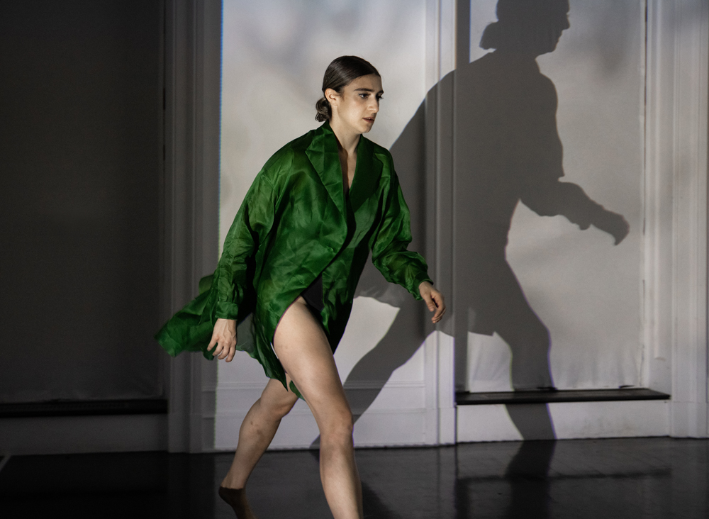 Wearing a green shirt dress, Xenia Mansour walks in profile. Her shadow looms ominously on he white wall behind her.